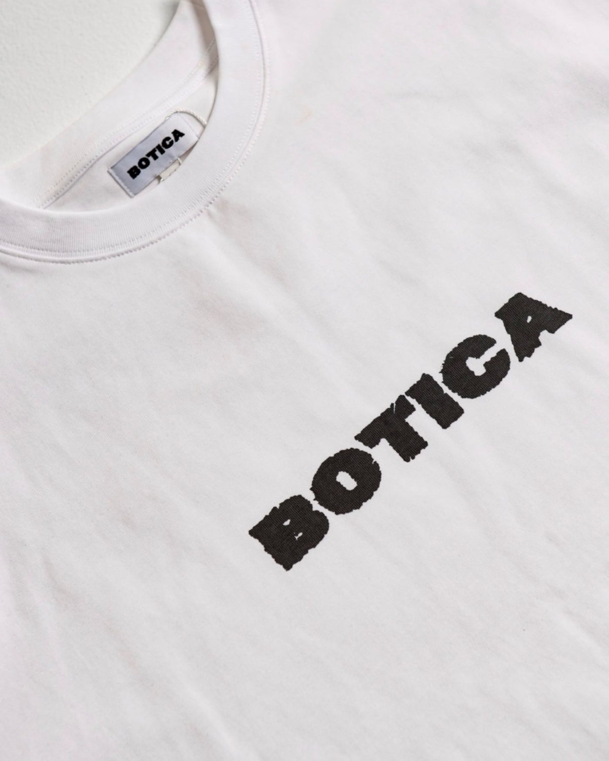 Close-up of the black BOTICA chest print and woven neck label on The Front Hit Tee in white