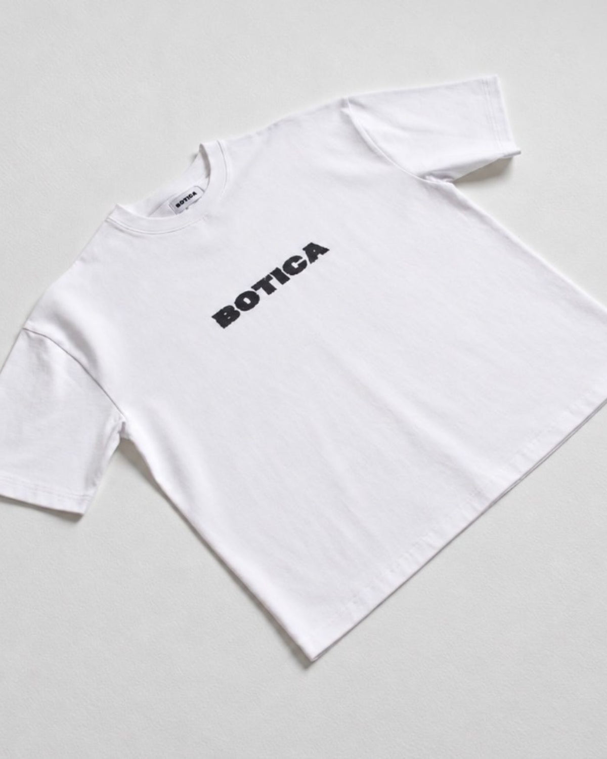 Flat lay of The Front Hit Tee in white with a black BOTICA print on the chest, on a pale grey background
