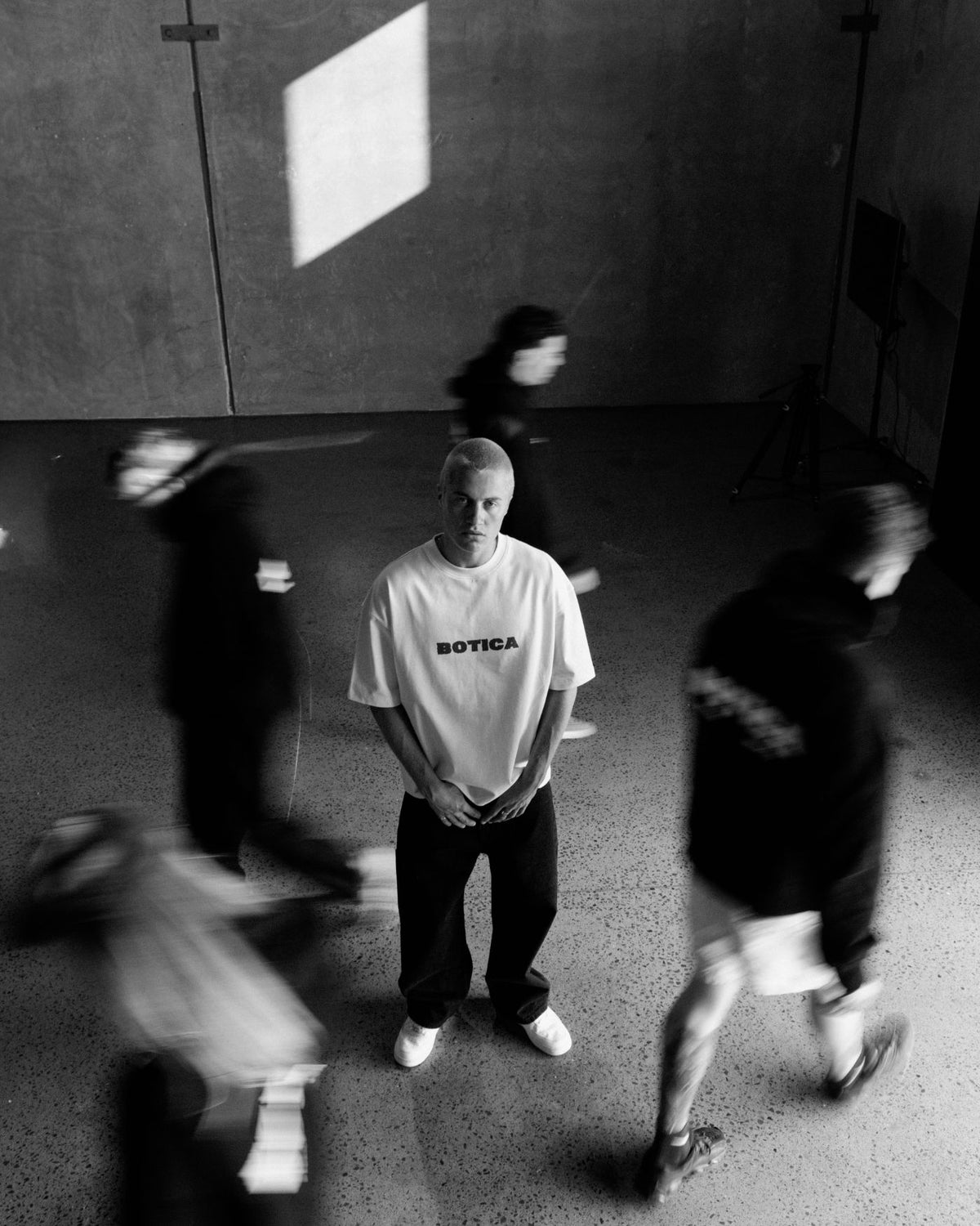 Model standing still in The Front Hit Tee in white and black trousers while blurred figures in black walk around them in a concrete studio