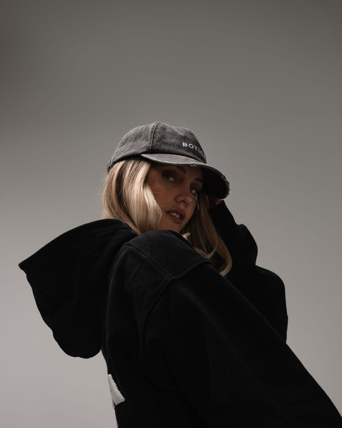 Model in the black Legacy Hoodie with the hood down and a washed grey BOTICA cap, looking over their shoulder