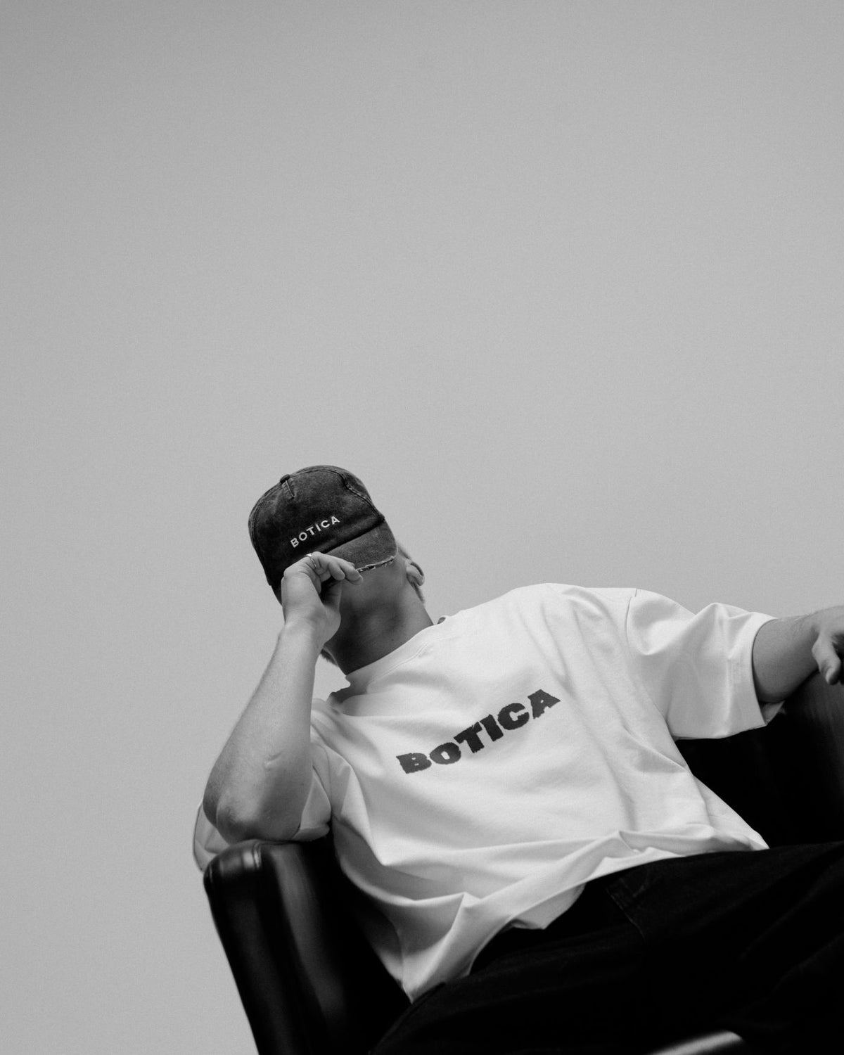 Model in a cap and The Front Hit Tee in white with a black BOTICA chest print, seated in a black leather armchair resting their head on one hand
