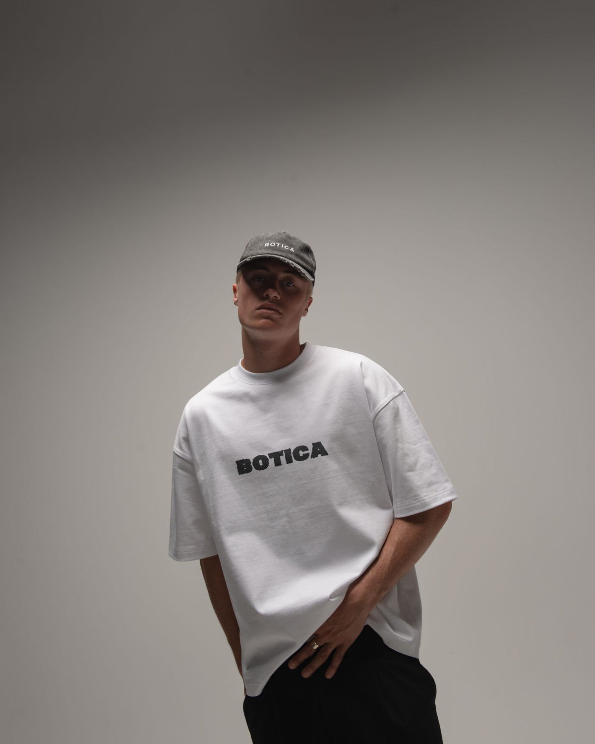 Model in a grey cap and The Front Hit Tee in white with a black BOTICA chest print, standing with one hand in a pocket against a grey studio backdrop