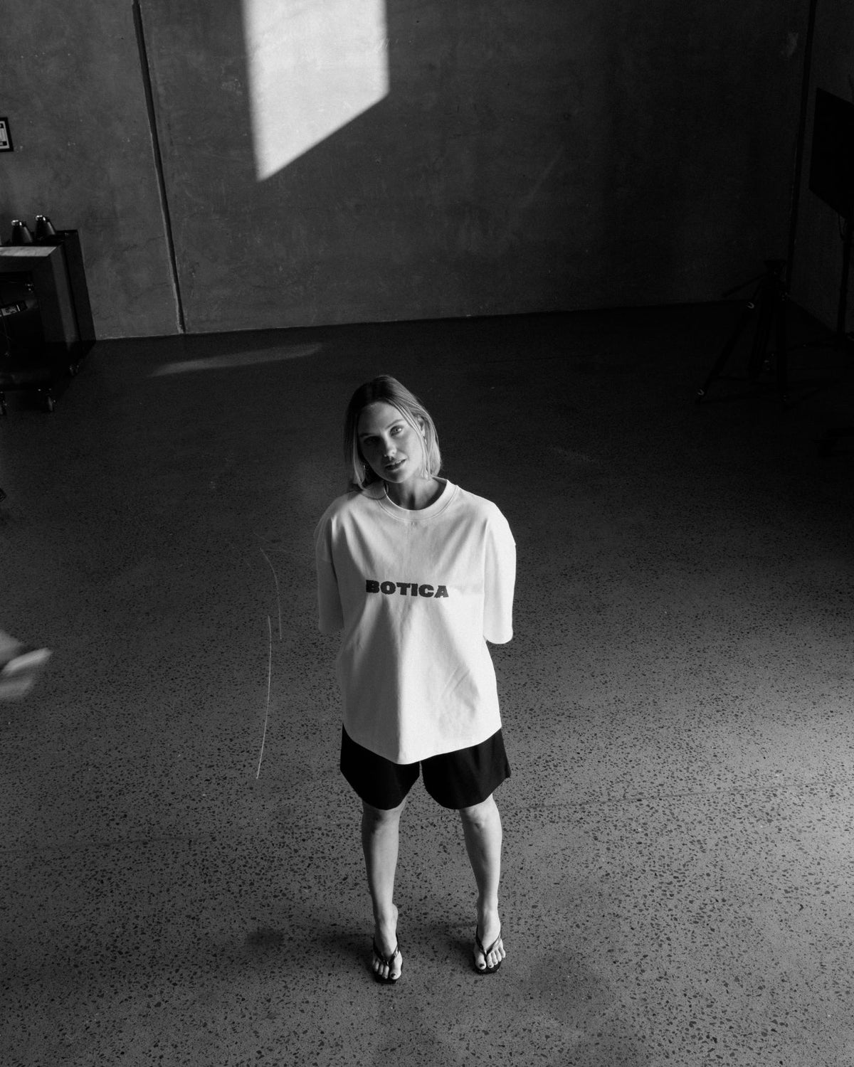 High-angle black and white photo of a model standing in a concrete studio wearing The Front Hit Tee in white with a black BOTICA chest print, black shorts and sandals