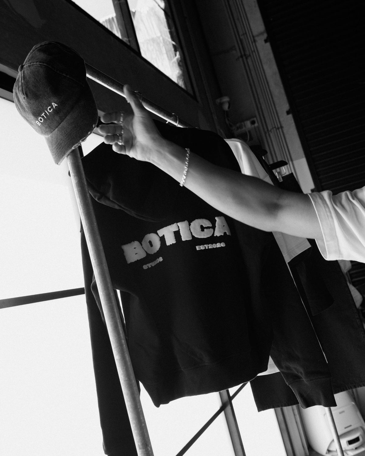 Model reaching for Botica cap and black Legacy Hoodie leaning on a metal frame, back print visible