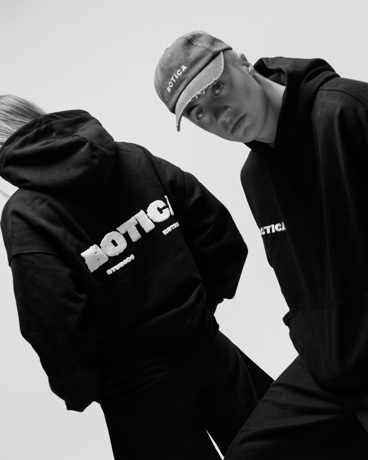 Black and white photo of two models in black Legacy Hoodies at a tilted angle against a pale studio backdrop, one with their back to the camera showing the raised BOTICA STUDIOS back print, the other facing forward in a washed BOTICA cap