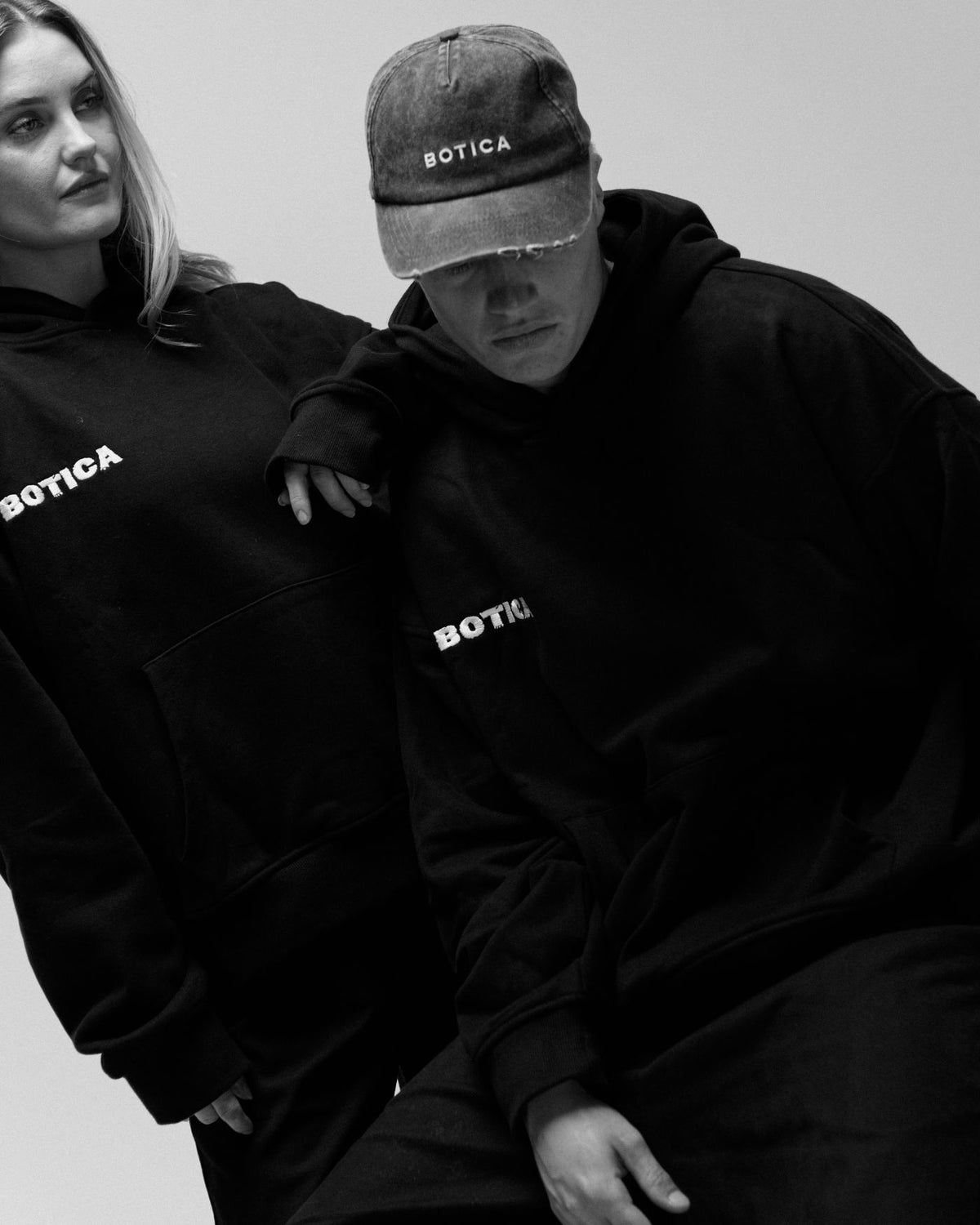 Two models in black Legacy Hoodies with the BOTICA chest logo, one wearing a washed grey BOTICA cap