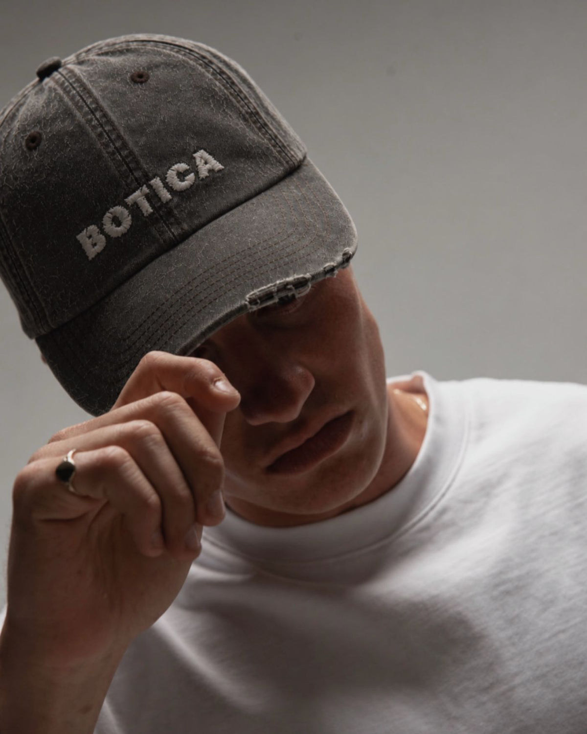 Model in The Logo Cap in washed black and a white tee, holding the peak with one hand, embroidered BOTICA wordmark visible