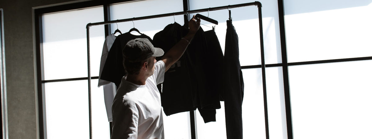 Man in a washed black cap and white tee reaching for a garment on a black metal clothing rail in front of large frosted windows