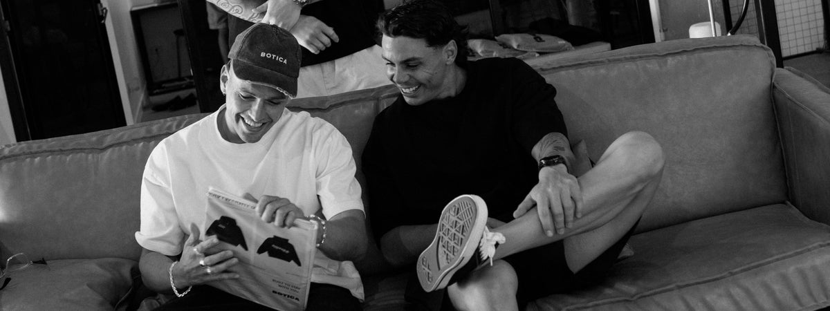 Black and white photo of two men laughing together on a couch while looking through a printed booklet of hoodie designs, one in a washed BOTICA cap and white tee, the other in a black tee