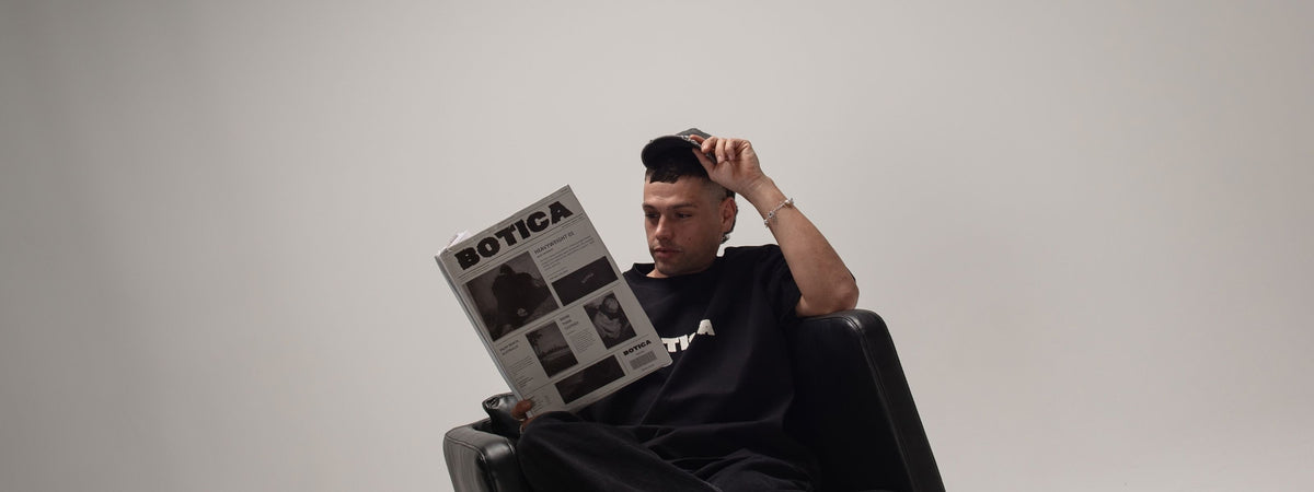 Man in dark clothing and a cap sitting in a black leather armchair, reading a printed BOTICA newspaper-style publication titled Heavyweight 01